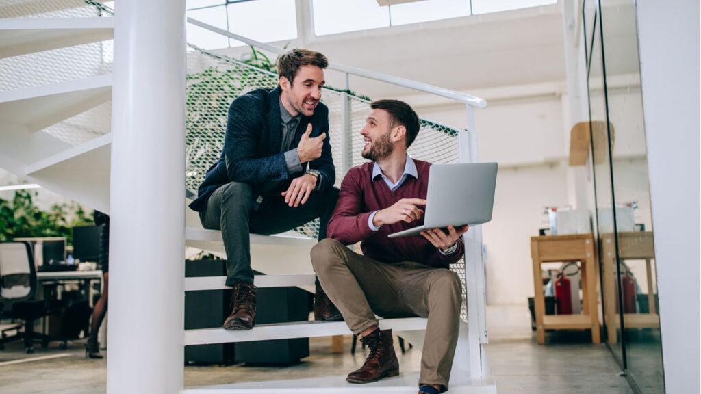 Zwei Geschäftsleute diskutieren ein Projekt auf einer Treppe im Büro, einer hält einen Laptop und beide lächeln.