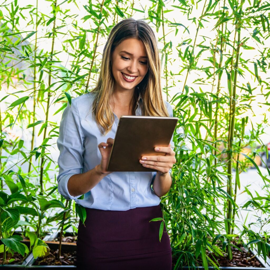 Geschäftsfrau arbeitet lächelnd an einem Tablet, umgeben von grünen Pflanzen in einem modernen Büro.